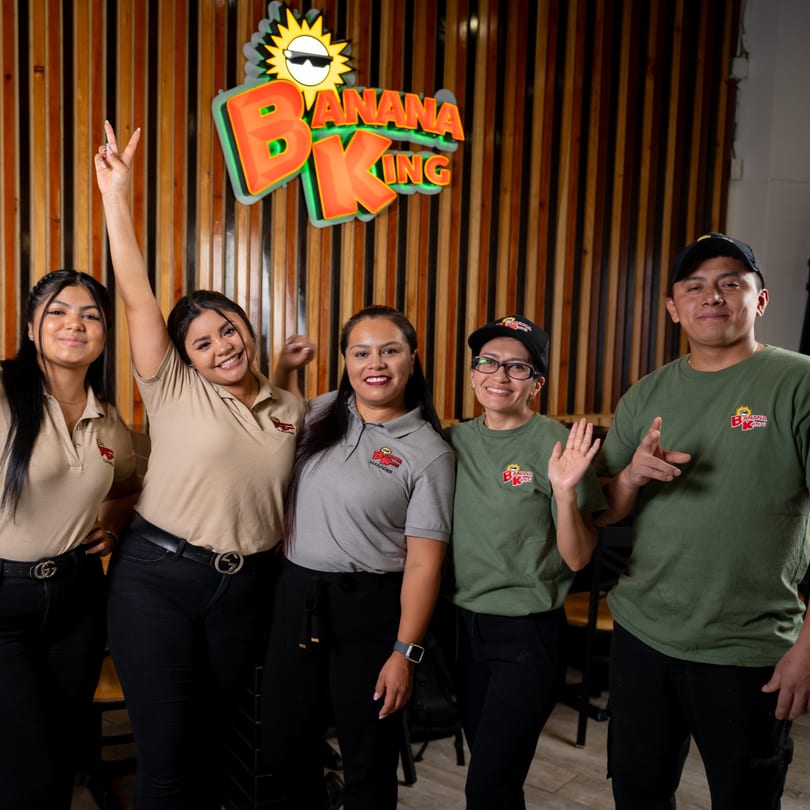 Five Banana King restaurant staff members, smiling and posing in front of a wooden wall with the company logo.