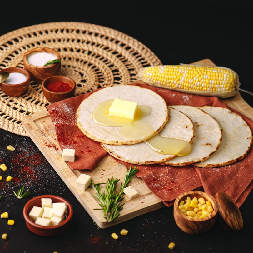 Corn tortillas with butter and honey, corn on the cob, spices, and cheese cubes on a dark background.
