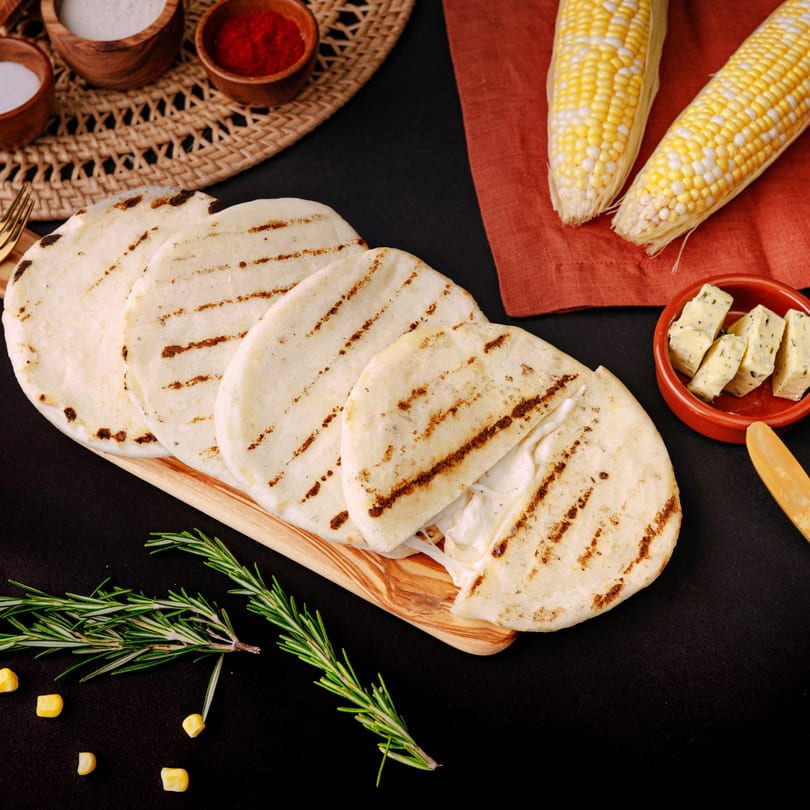 Grilled arepas with melted cheese, corn, rosemary, and spices on a dark surface.
