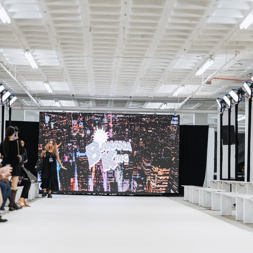 A woman in a black dress walks a runway with the Banana King logo on a large screen.