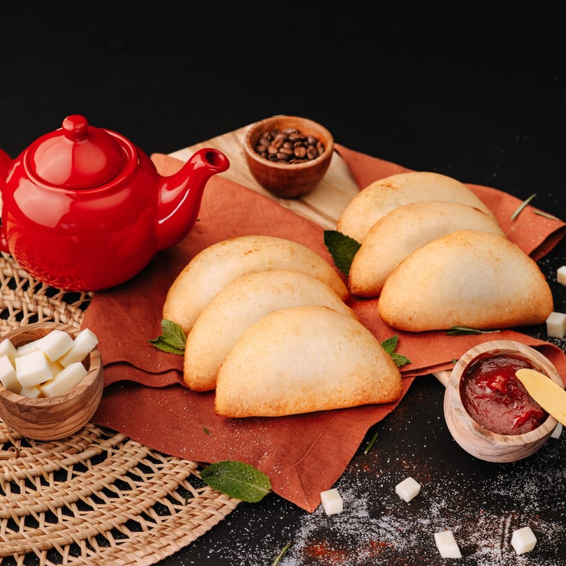 Golden empanadas on an orange napkin with a red teapot, coffee beans, sugar, and jam.