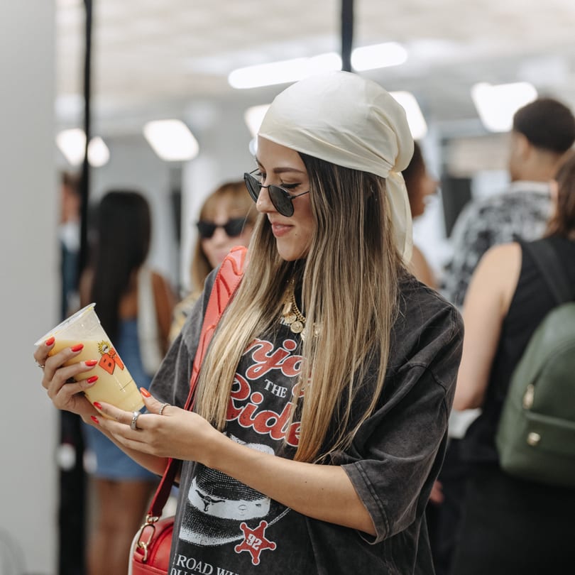 A young woman in sunglasses and a headscarf smiles while holding a Banana King smoothie.