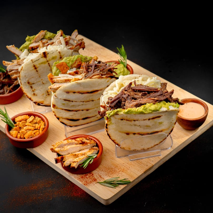 Three arepas filled with various meats, guacamole, and cheese, served on a wooden board.