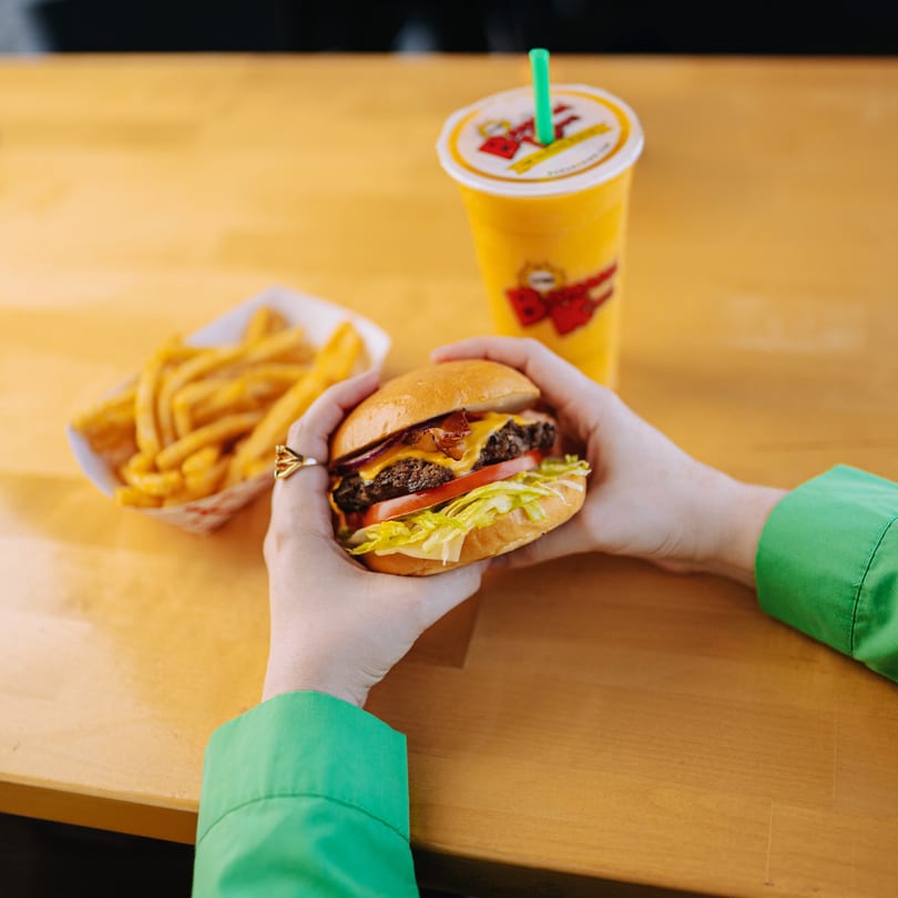 Hands holding a Banana King cheeseburger with fries and a drink on a wooden table.