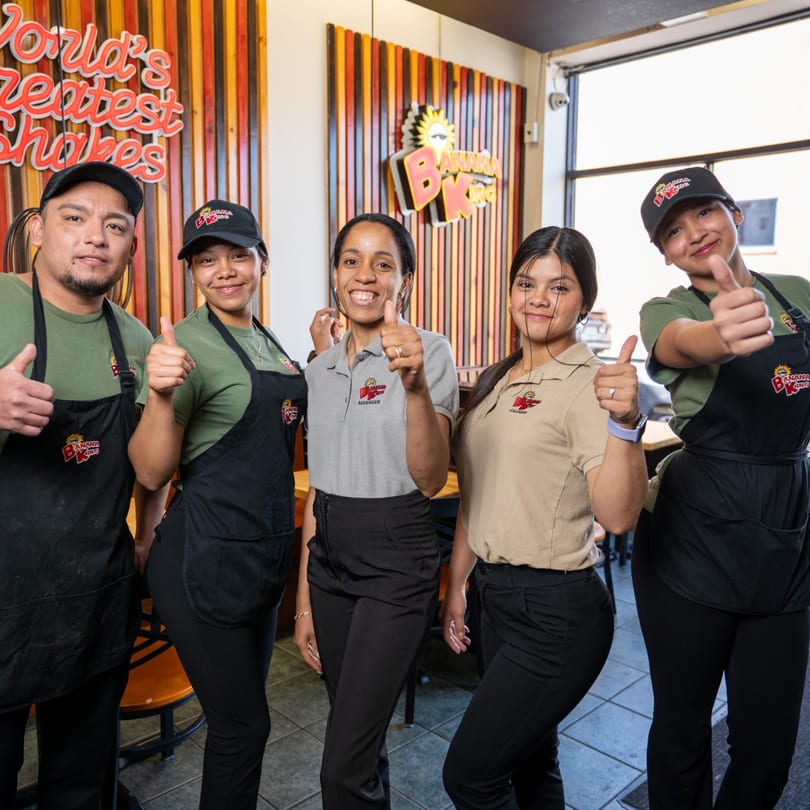 Five Banana King employees, including a manager and cashier, smiling and giving thumbs up in the restaurant.