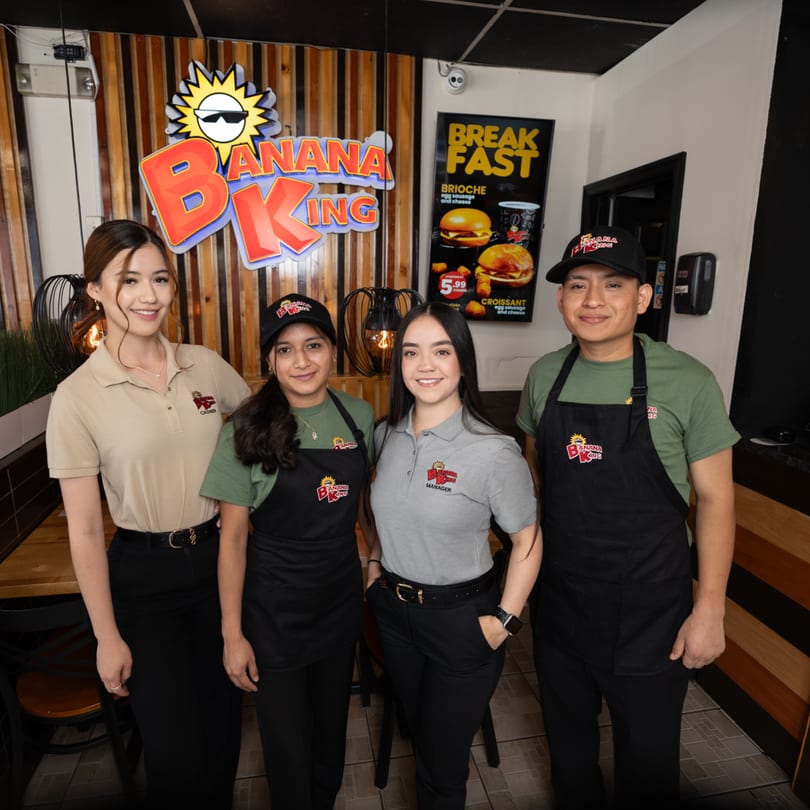 Four Banana King employees, two women and two men, stand smiling in front of the restaurant's logo and breakfast menu.