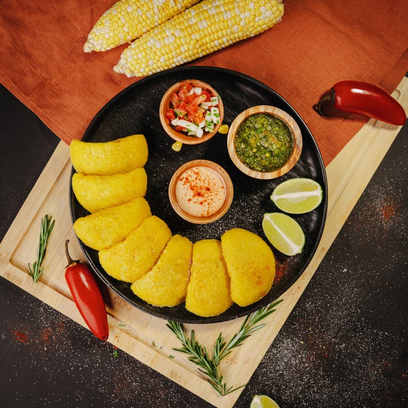 A black plate of golden empanadas with three dipping sauces, limes, and corn on the cob.