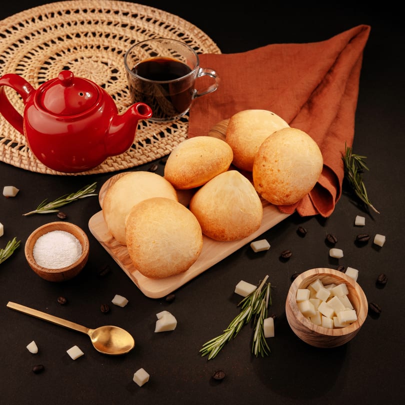Golden cheese bread rolls, a red teapot, and coffee on a dark table with sugar and rosemary.