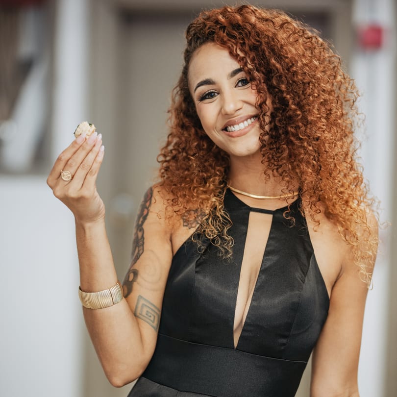 Smiling woman with curly red hair holding a small dessert, wearing a black dress.