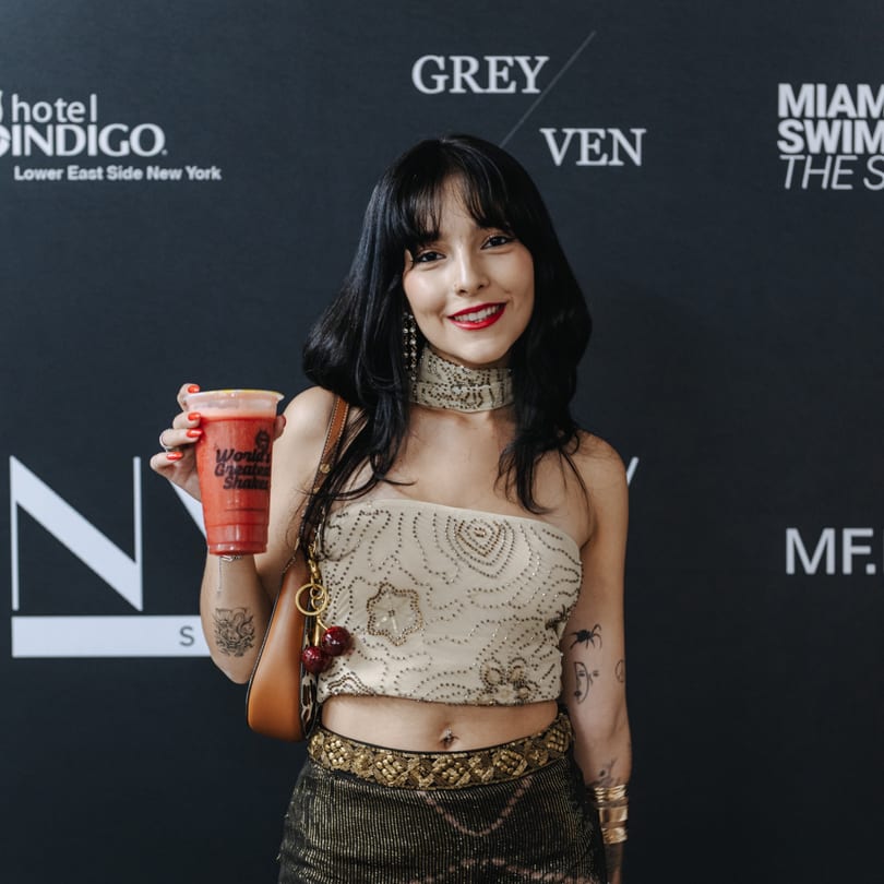 A smiling woman in a sequined top holds a red Banana King shake against a dark branded background.
