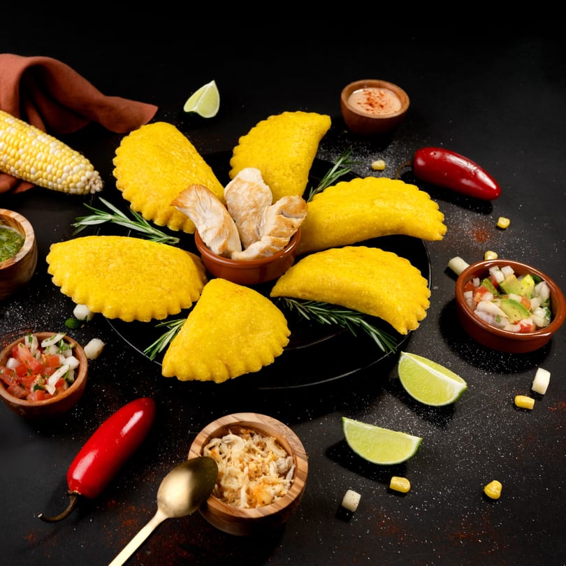 Golden empanadas on a black plate, surrounded by corn, peppers, limes, and various dips.